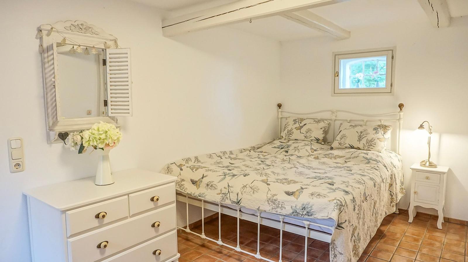 Schlafzimmer mit weißem Bett, Schrank und Nachttisch. Holzbalken und Fenster sichtbar.