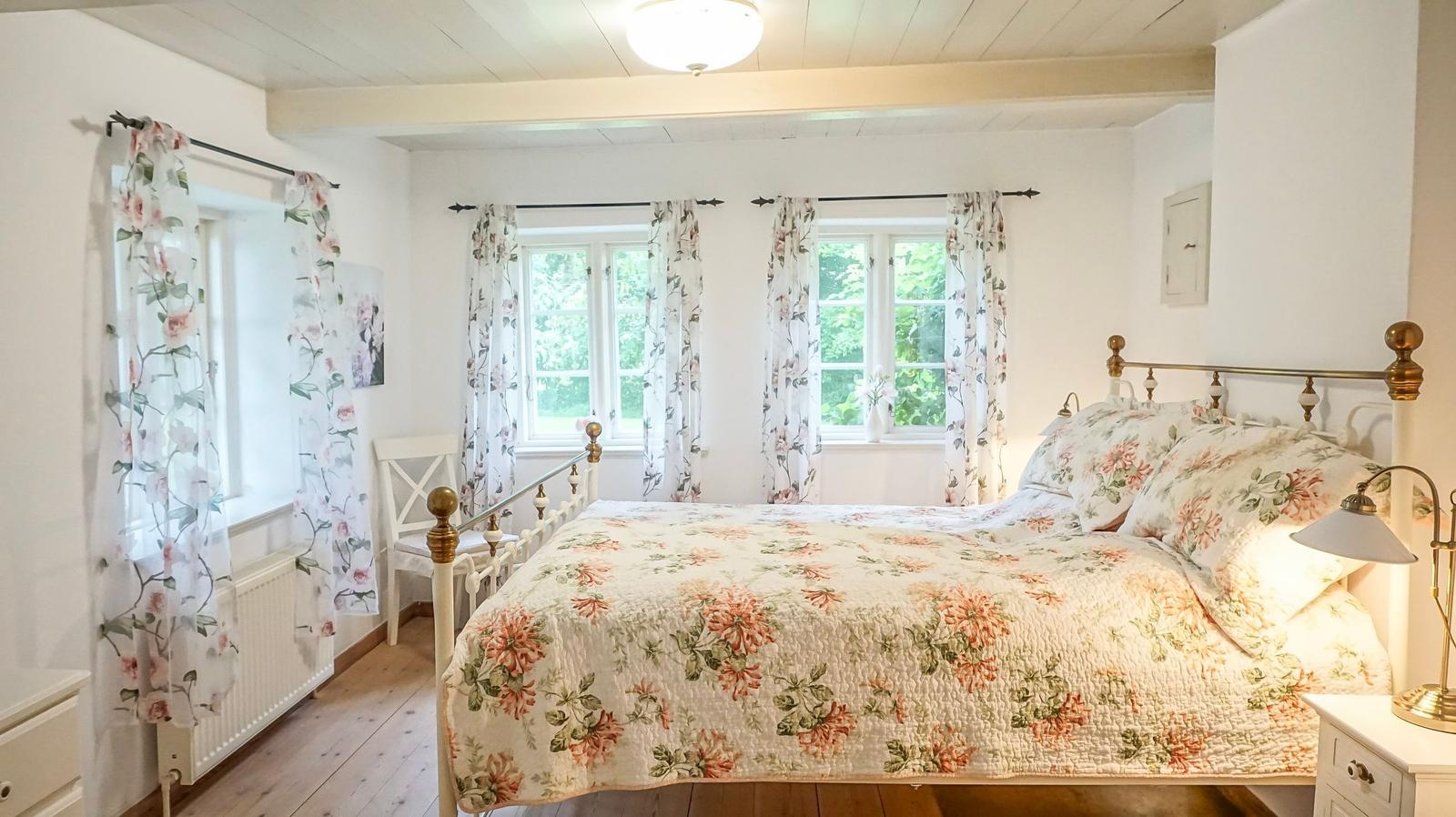 Schlafzimmer mit Bett, Fenstern und floralen Vorhängen.