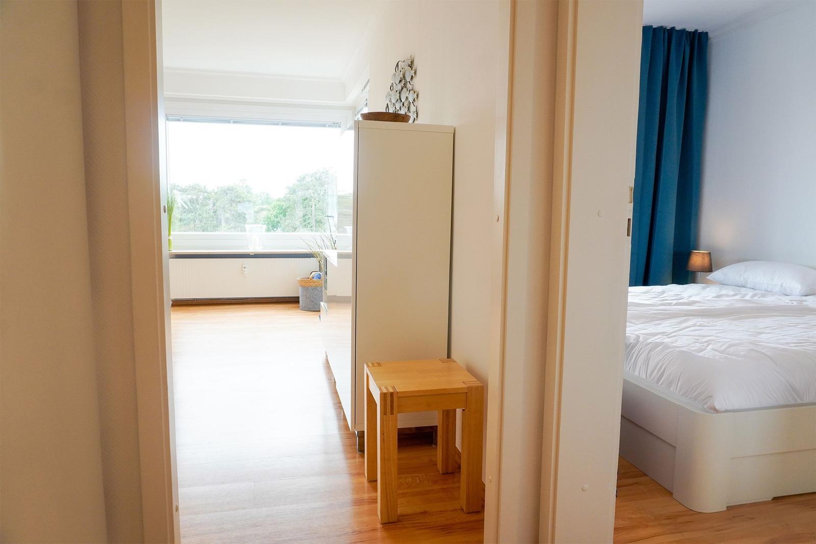 Schlafzimmer mit Bett und blauen Vorhängen. Durchgang zu Wohnbereich mit Fenster und Holzboden.