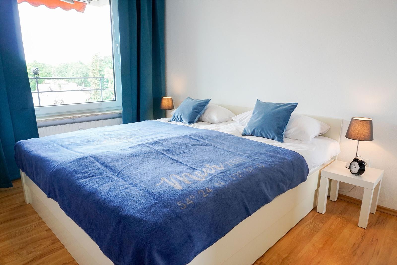 Schlafzimmer mit Bett, blauer Tagesdecke und Fenster mit Blick.