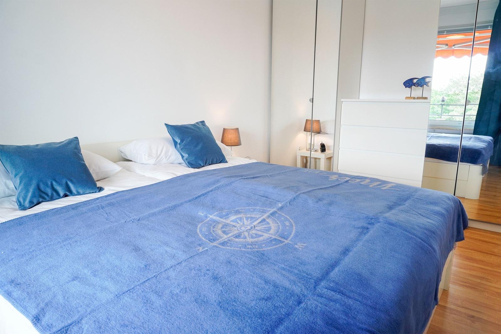 Schlafzimmer mit Bett, blauer Decke und Spiegelschrank. Blick auf Balkon mit Blick auf Grün.