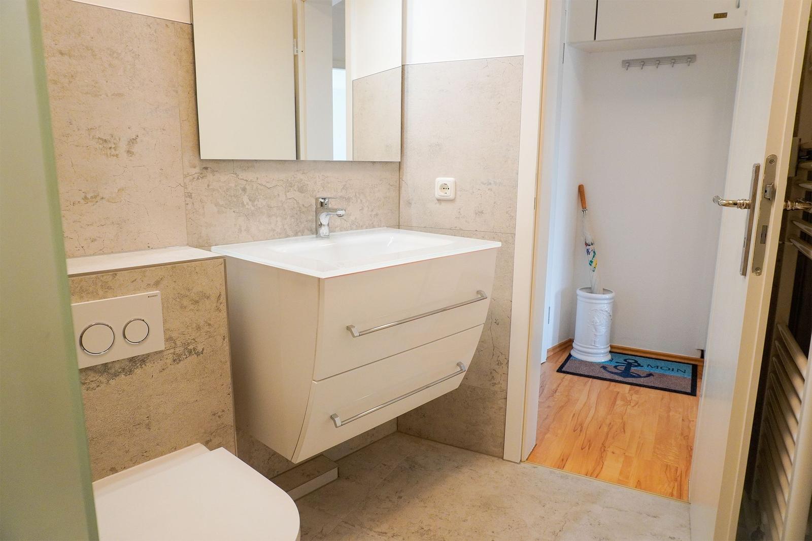 Badezimmer mit Waschbecken, Spiegelschrank und Toilette. Durchgang zu Flur mit Holzboden.