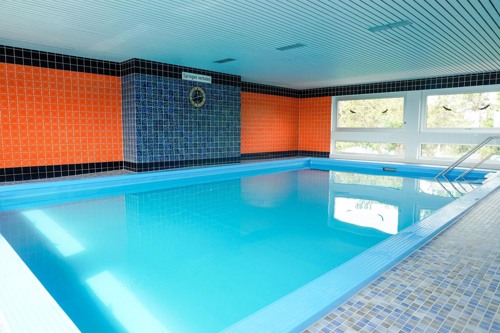 Innere Schwimmhalle mit blauem Wasser, orangefarbenen und blauen Fliesen an den Wänden und Fenstern zur Aussenseite.