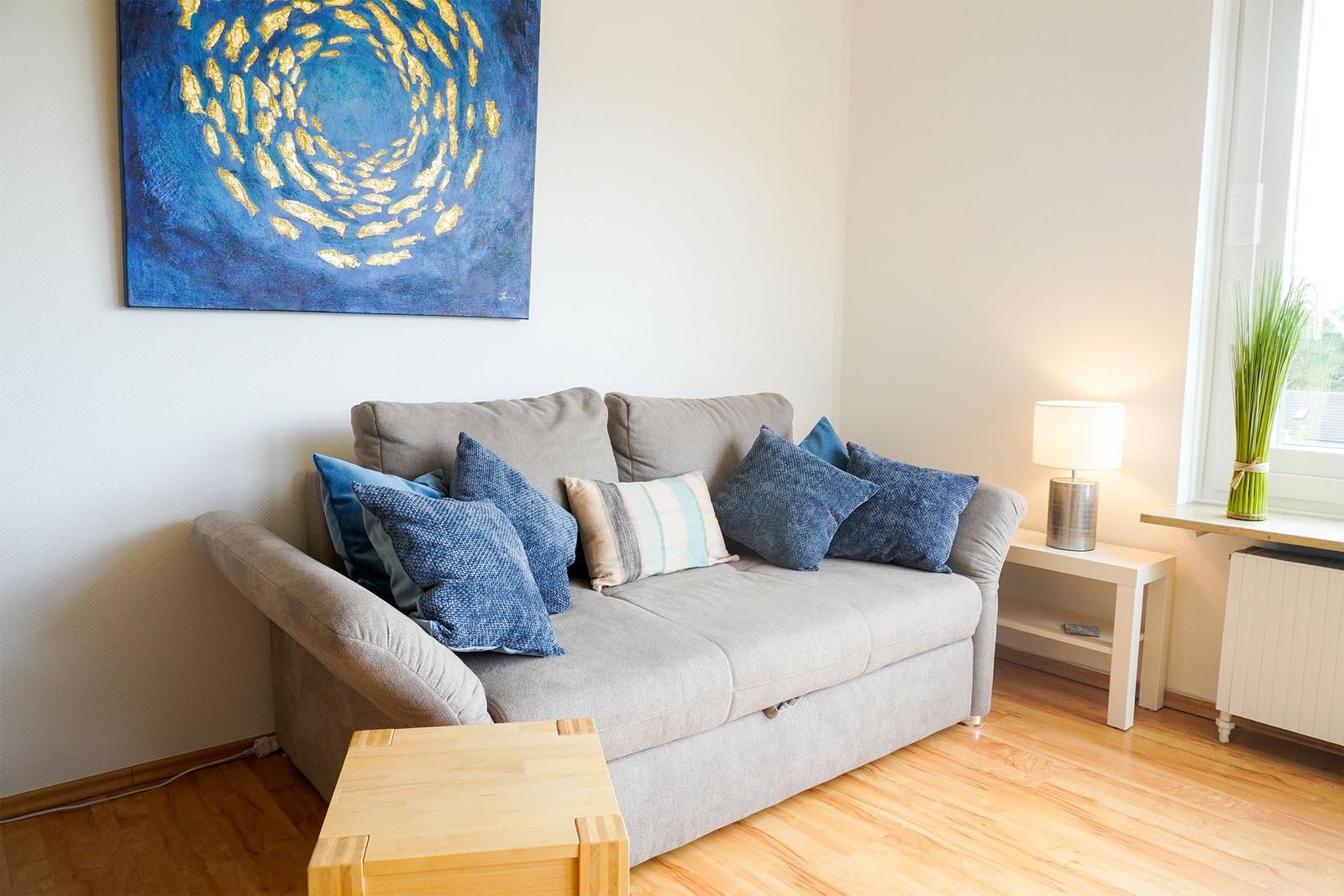Ein grauer Sofa mit blauen Kissen und einem Holztisch. An der Wand hängt ein blaues Gemälde.