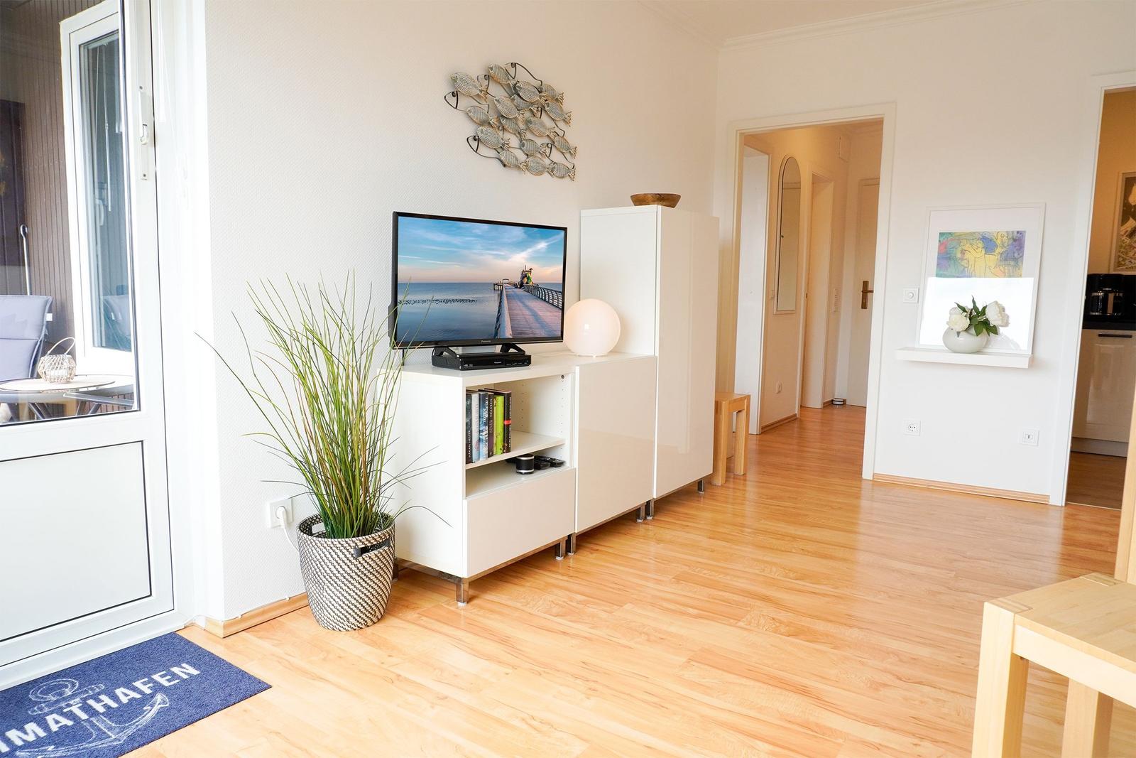 Wohnzimmer mit Fernseher, Pflanze und Holzboden. Tür führt zum Flur und zur Küche.