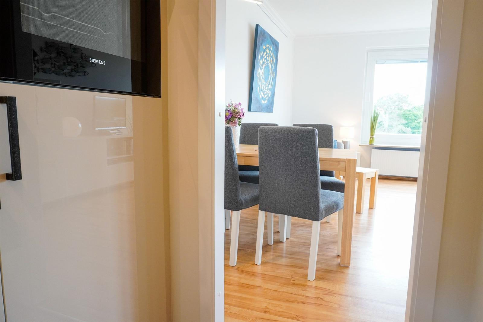 Esszimmer mit Holztisch, grauen Stühlen und Fensterblick. Küchenzeile mit integriertem Ofen.