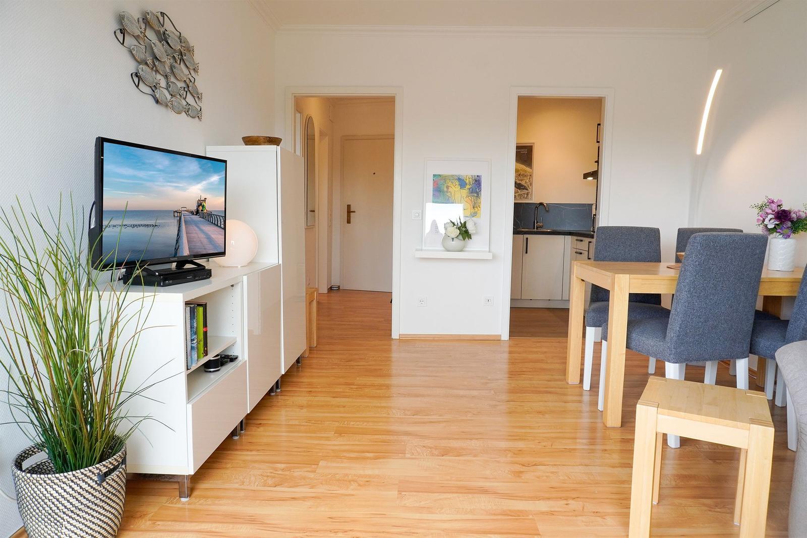 Wohnbereich mit TV, Esstisch und Küchenbereich. Holzboden und weiße Möbel.