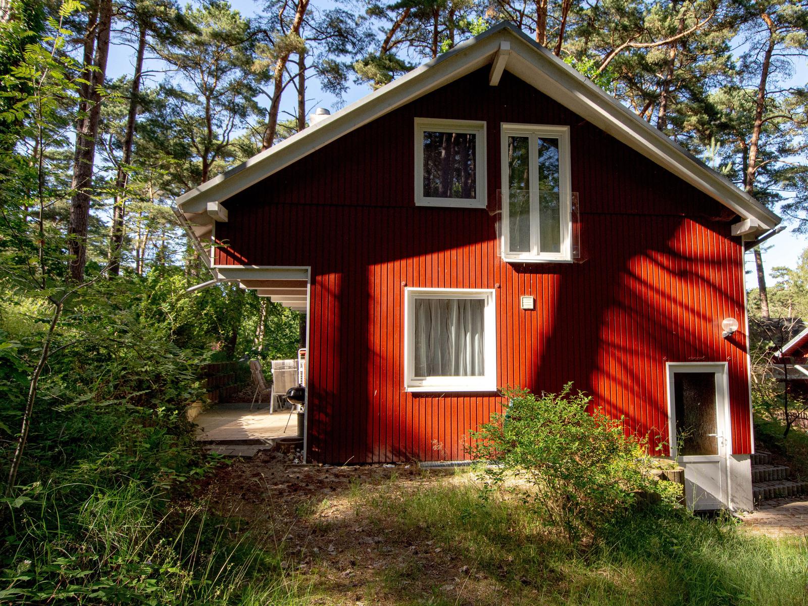 Rotes Holzhaus mit weißen Fenstern im Wald. Terrasse mit Sitzgelegenheit.