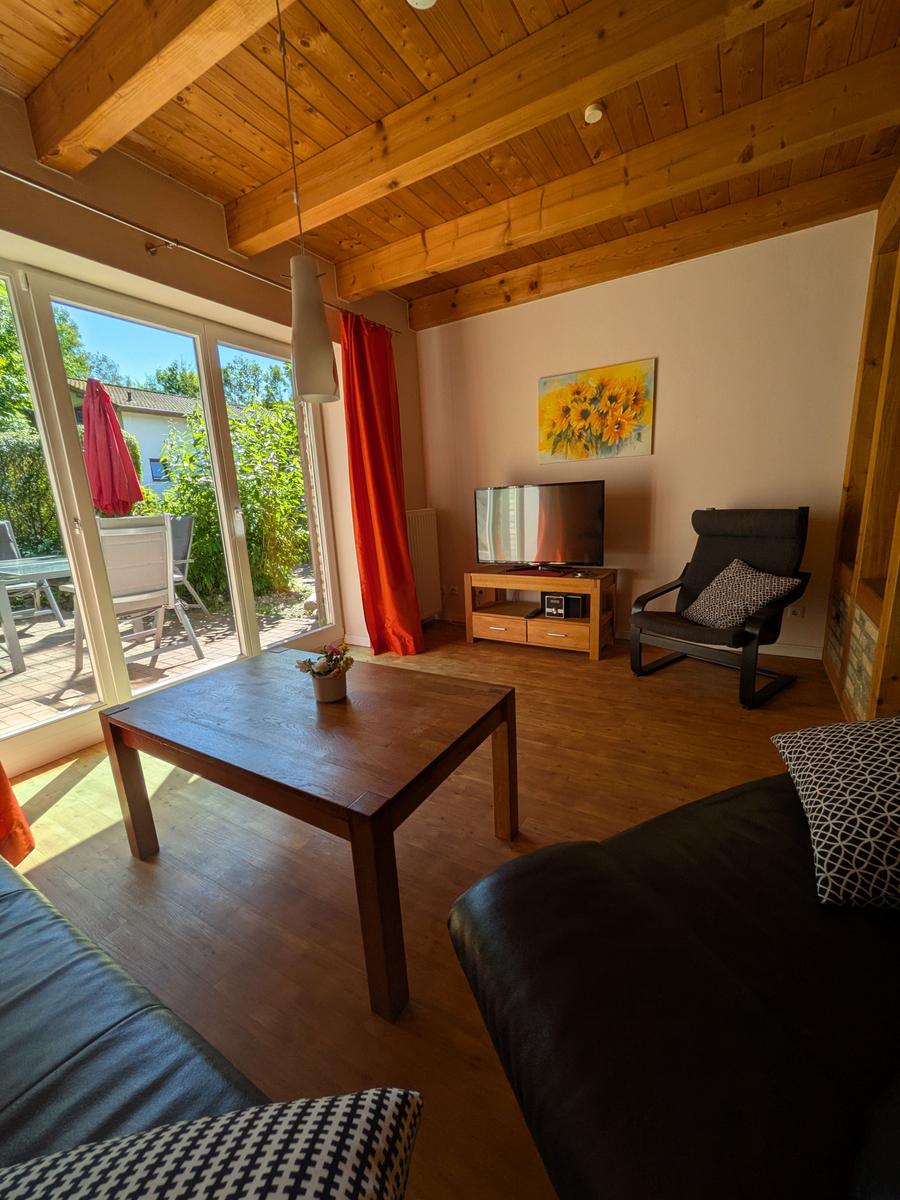 Wohnzimmer mit Holzdecke, Sofa, Tisch und TV. Durchgang zur Terrasse mit Garten.