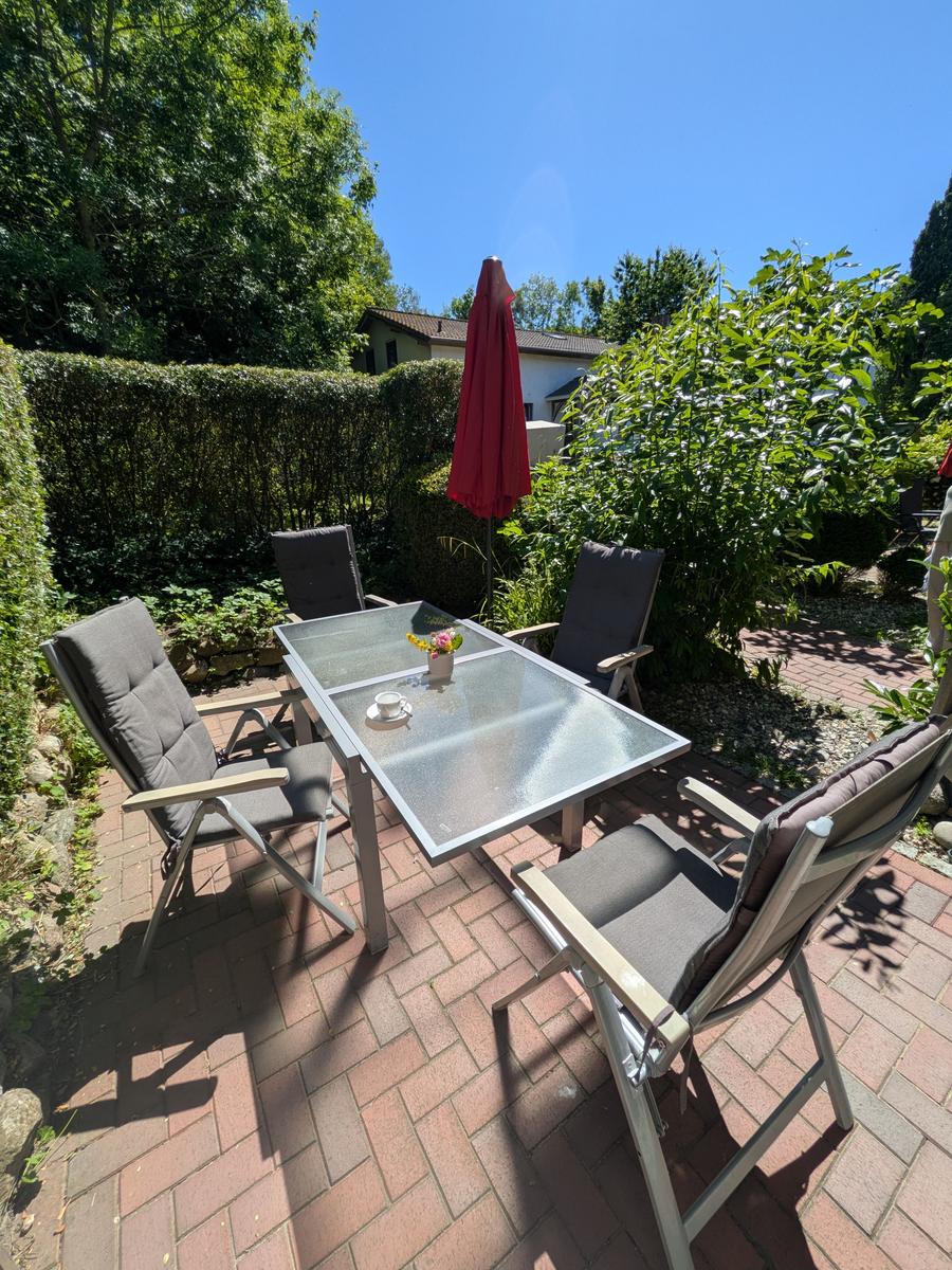 Terrasse mit Glas-Tisch, vier Stühlen und rotem Sonnenschirm. Umgeben von grüner Hecke und Bäumen.