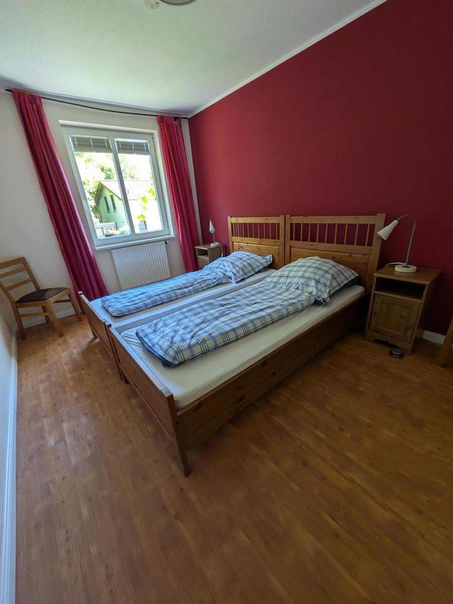 Zimmer mit Doppelbett, Holzboden und roter Wand. Fenster mit Vorhängen und Nachttische.