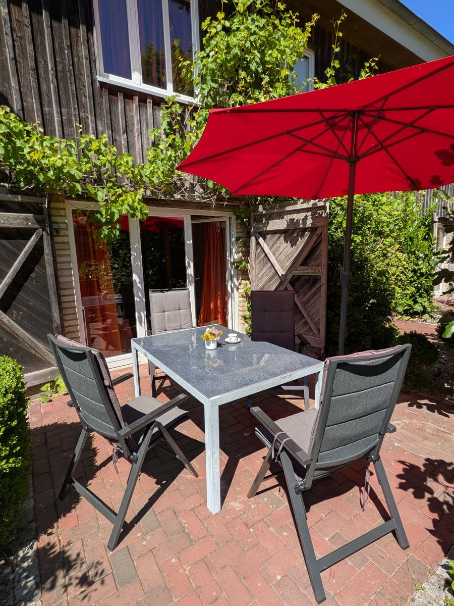 Terrasse mit Tisch, Stühlen und rotem Sonnenschirm vor Holzhaus mit Kletterpflanzen.