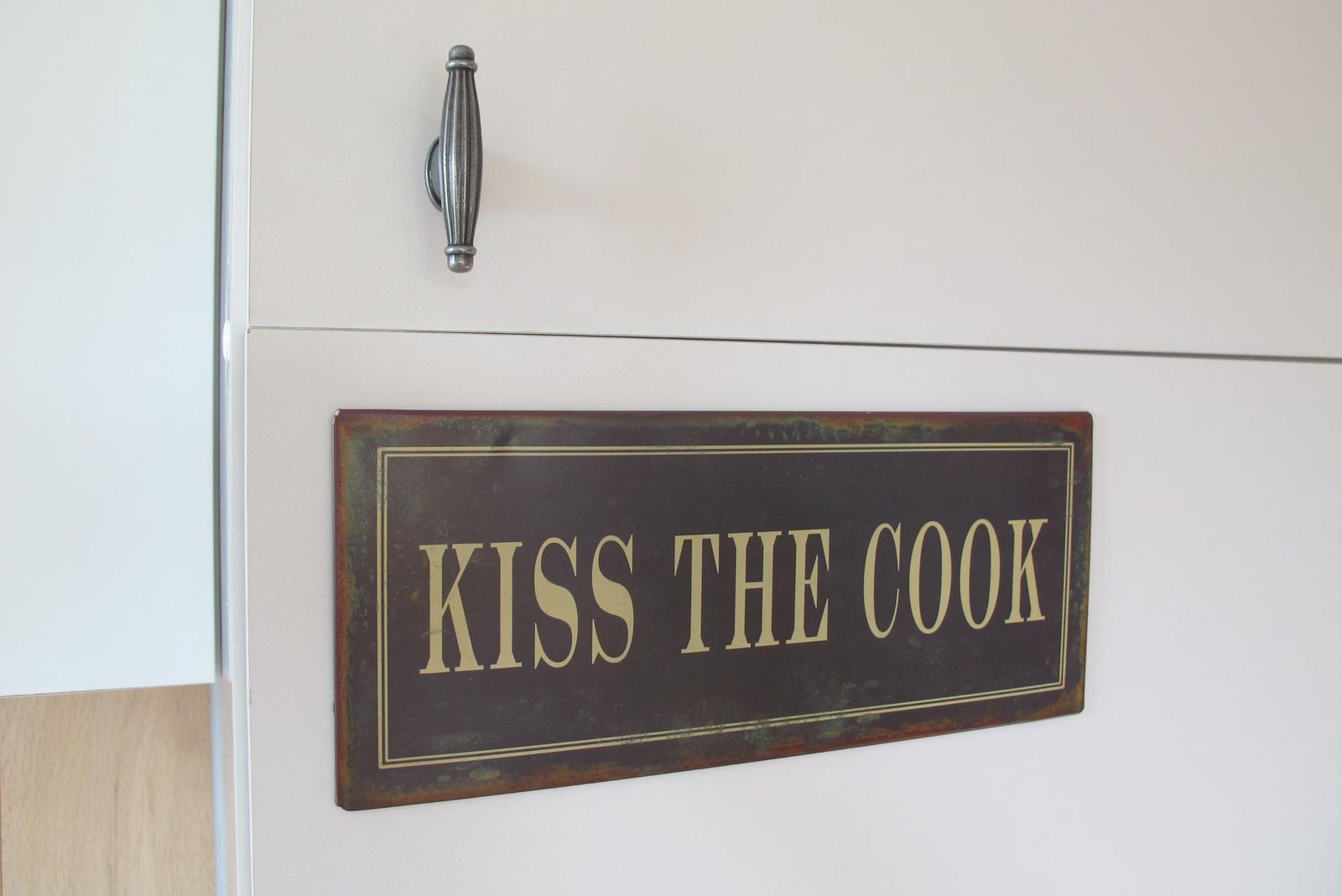 Schild mit der Aufschrift KISS THE COOK an einer Küchenwand.