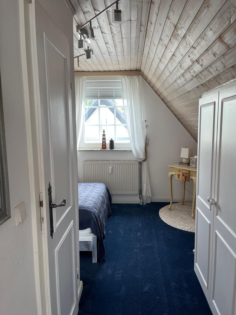 Schlafzimmer mit Dachschräge, blauem Teppich und Fenster mit Vorhängen.