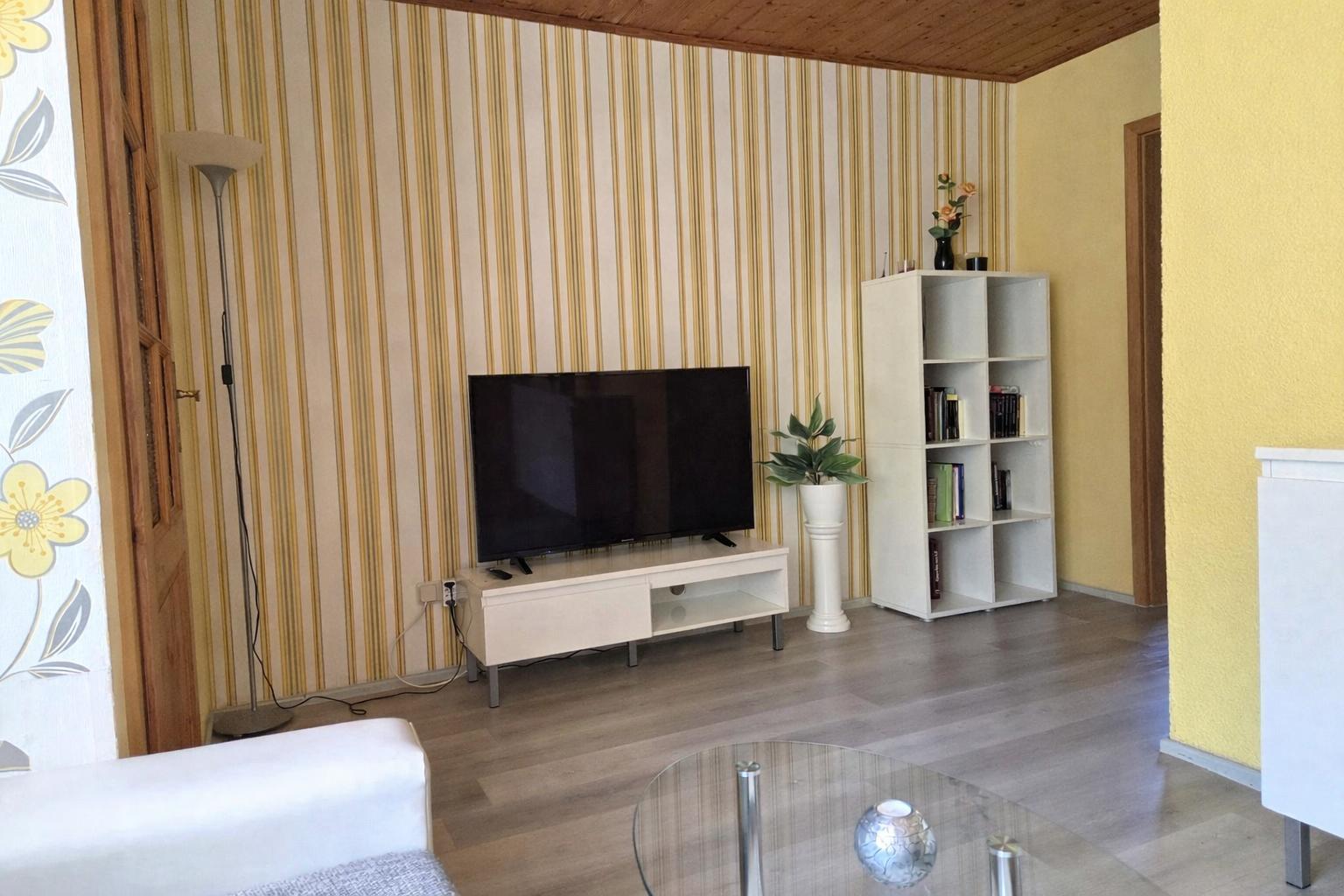 Wohnzimmer mit Fernseher, Bücherregal und Glas-Tisch.