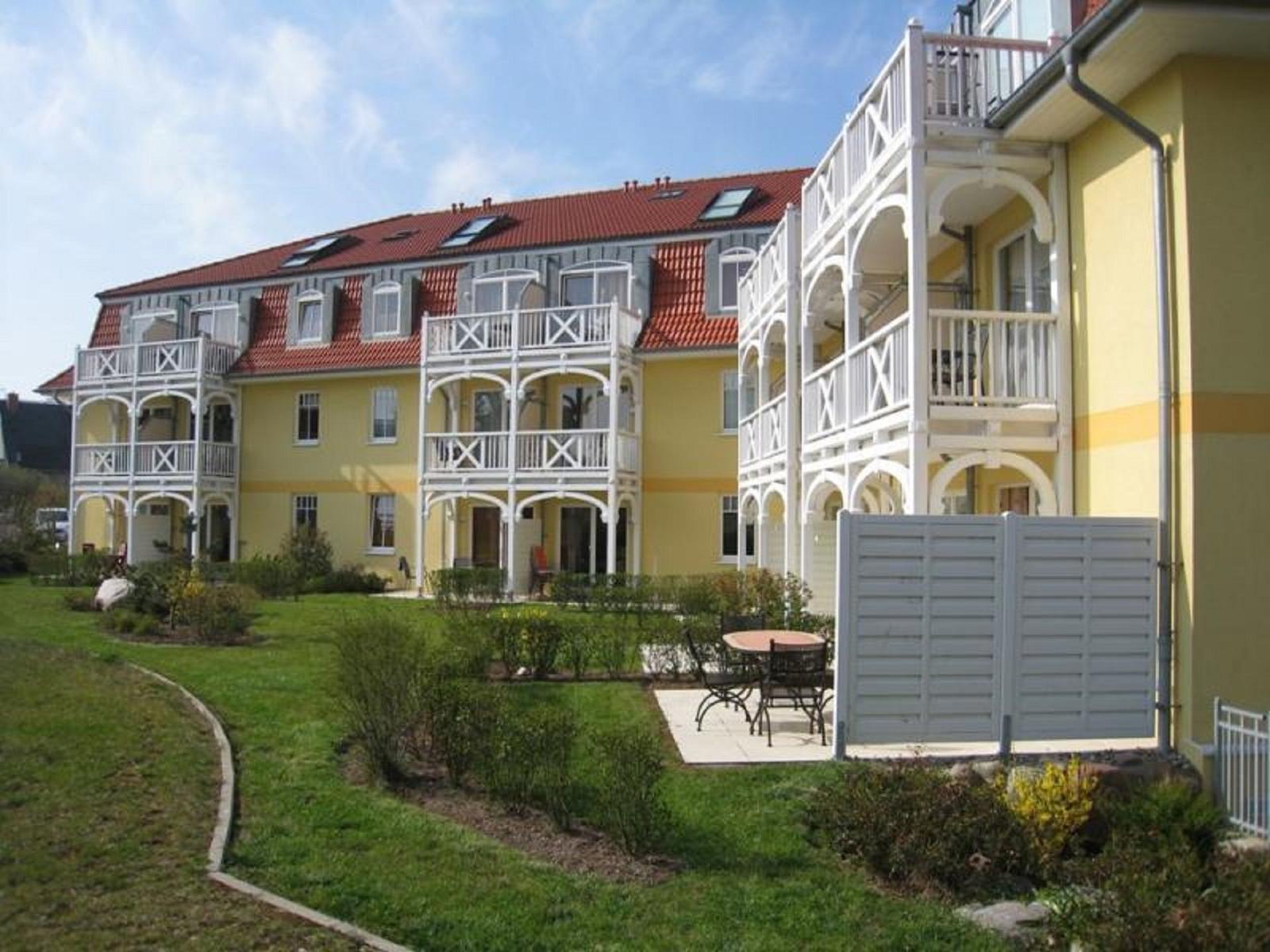 Gelbes Mehrfamilienhaus mit weißen Balkonen und rotem Dach. Gartenbereich mit Tisch und Stühlen.