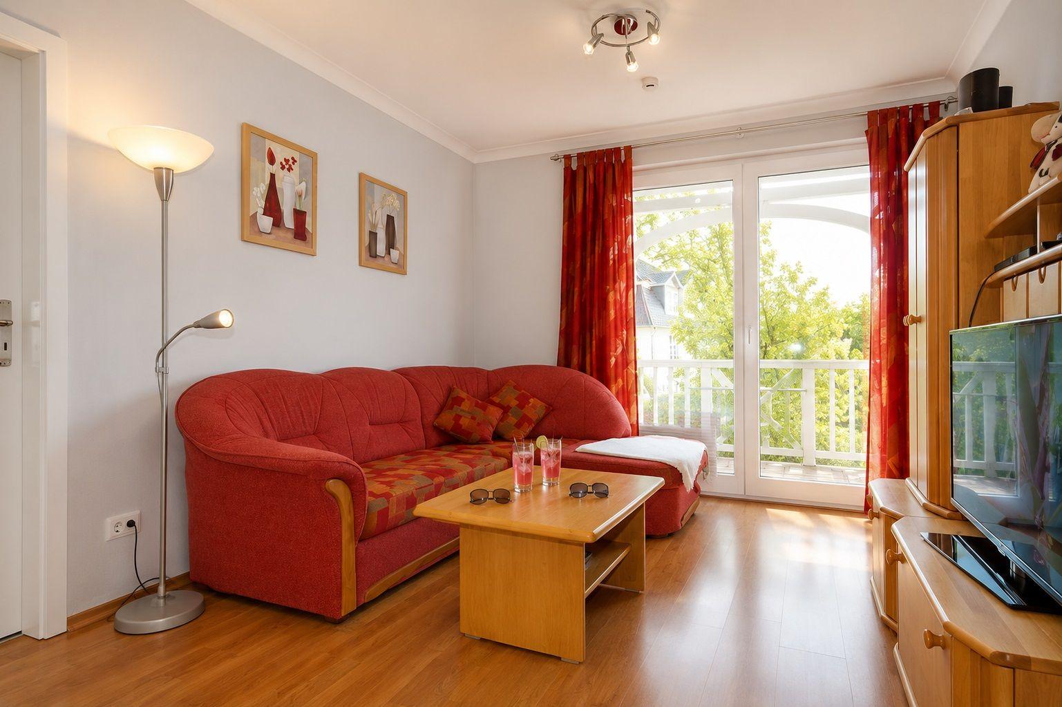 Ein Wohnzimmer mit rotem Sofa, Holztisch und TV. Durchgehende Glastüren führen auf eine Terrasse.