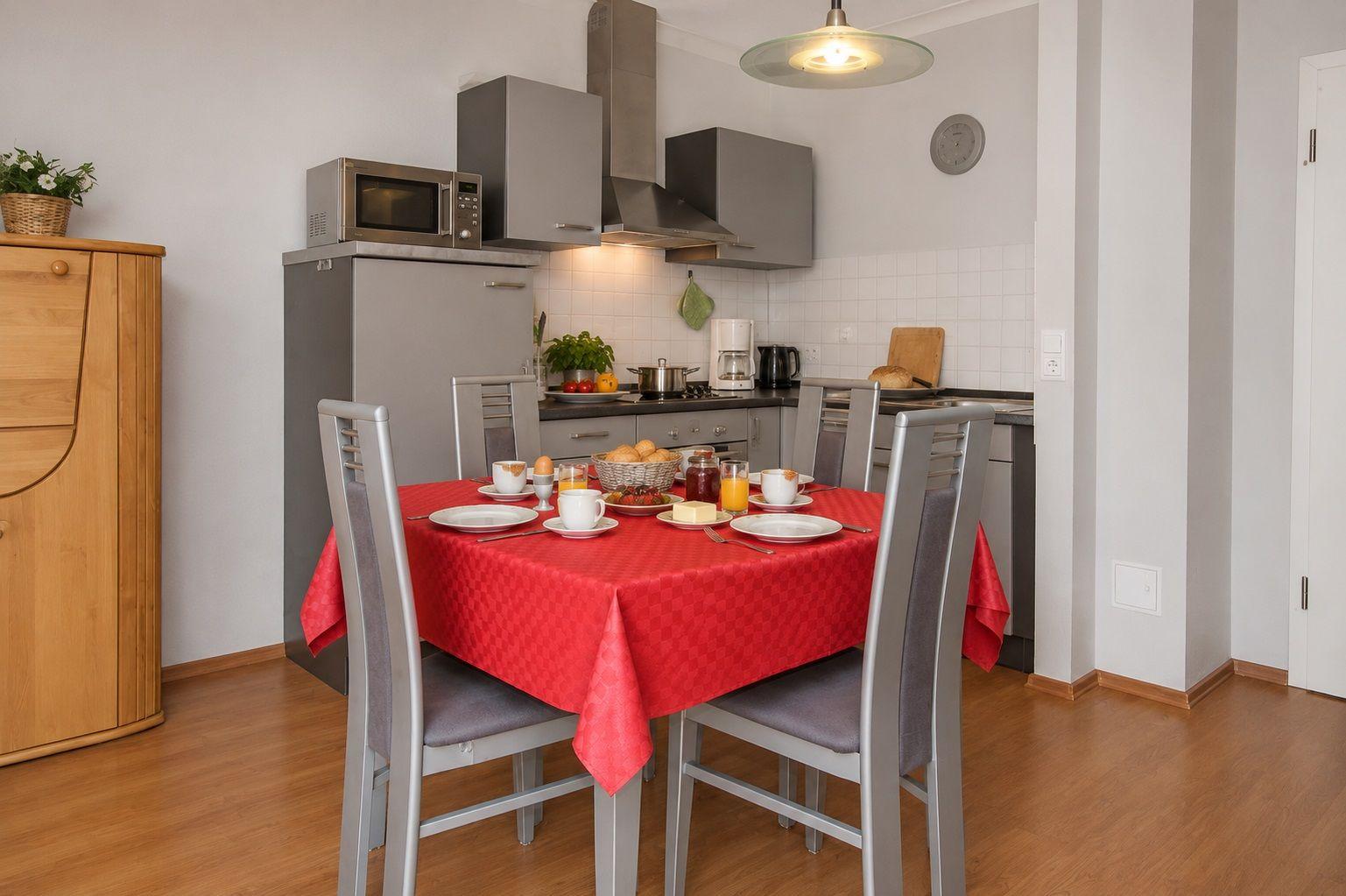 Küche mit Esstisch, roter Tischdecke und vier Stühlen. Hintergrund: Küchenschränke und Küchenzeile.