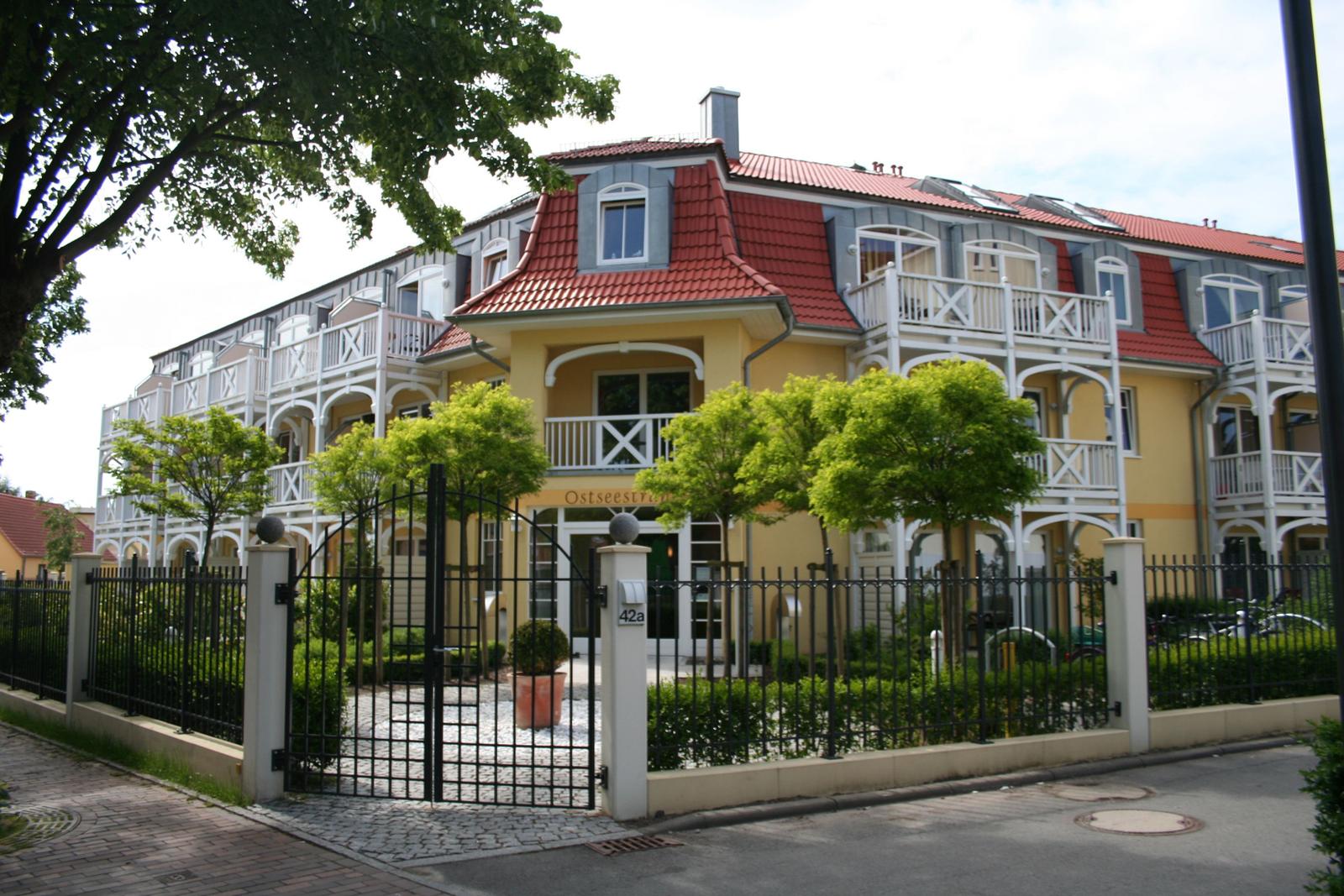 Haus mit rotem Dach und weißen Balkonen hinter schwarzem Zaun.