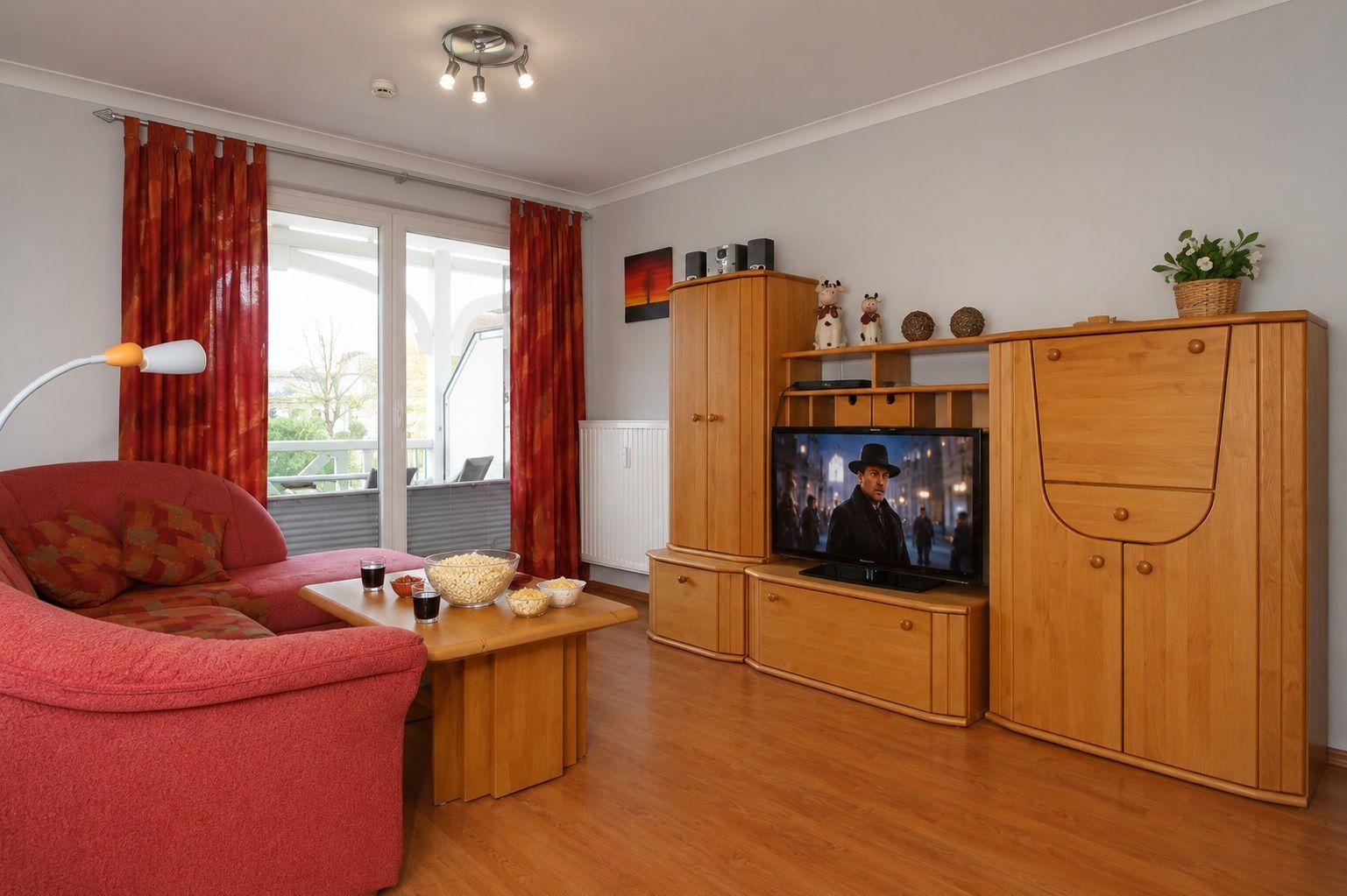 Ein Wohnzimmer mit rotem Sofa, Holzmöbeln und Fernseher. Durchgang zur Terrasse mit roten Vorhängen.