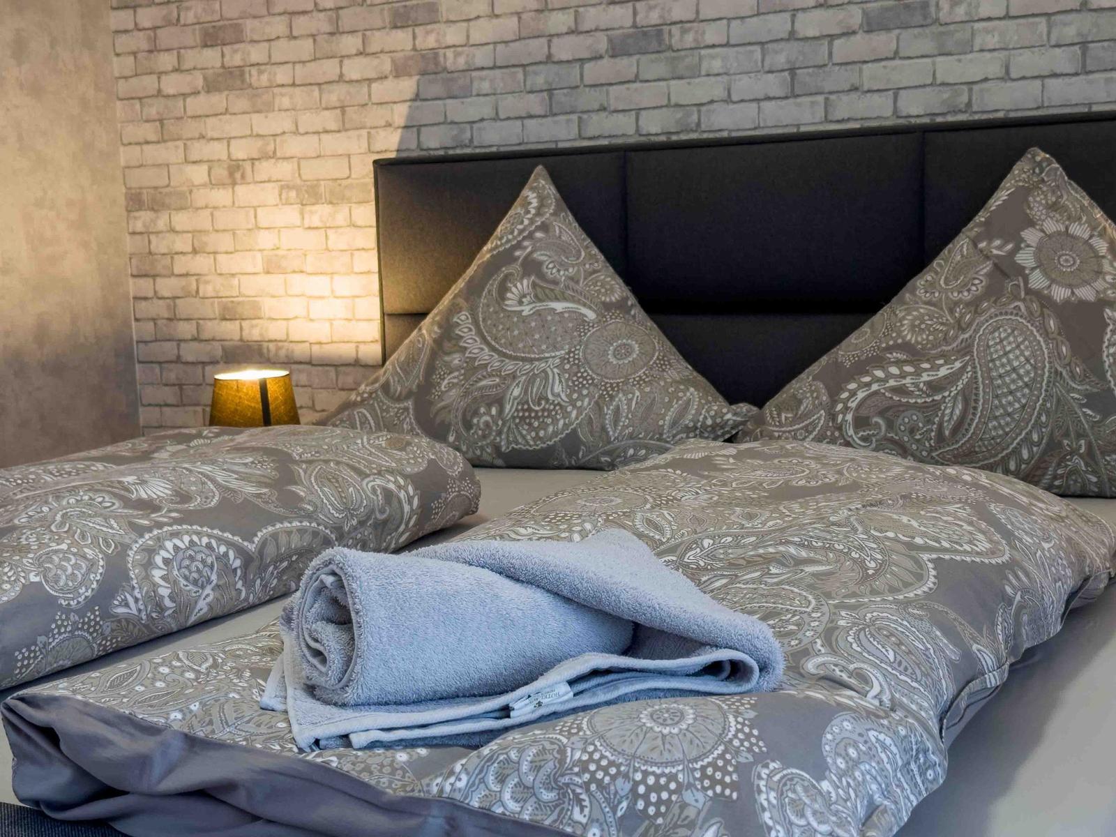 Ein Bett mit gemustertem Bettzeug und einem Handtuch darauf. Hinter dem Bett ist eine Ziegelwand.