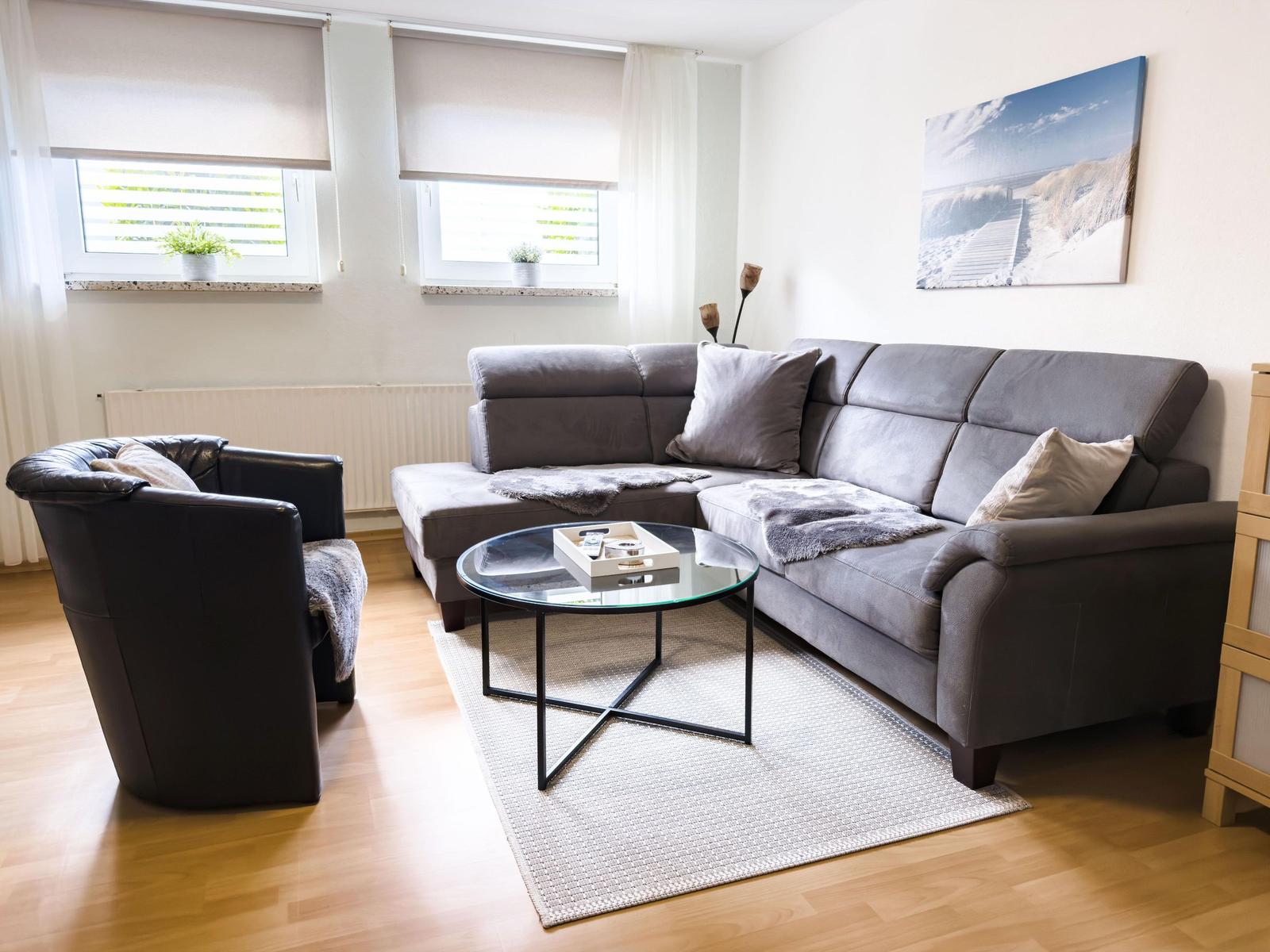 Ein Wohnzimmer mit grauem Sofa, Glas-Tisch und zwei Fenstern mit Pflanzen.