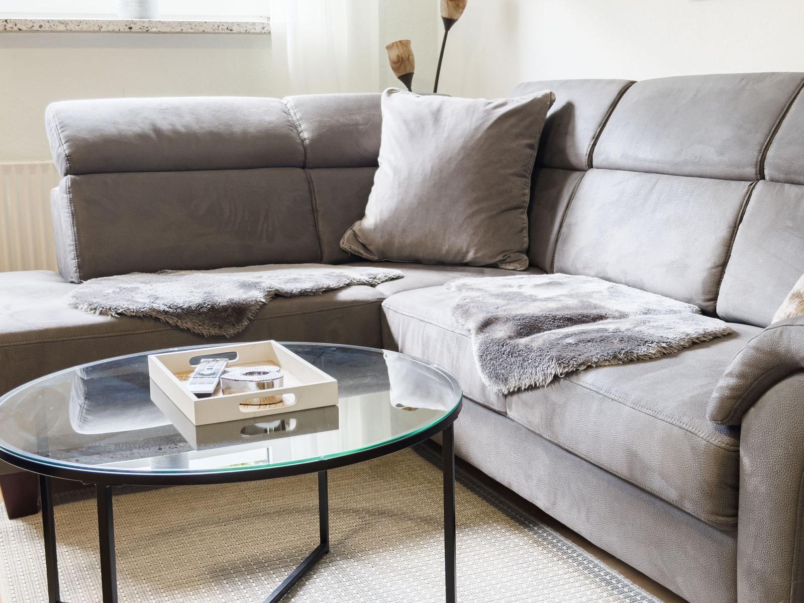 Ein grauer Sofa mit Kissen und Decke vor einem Glas-Tisch.