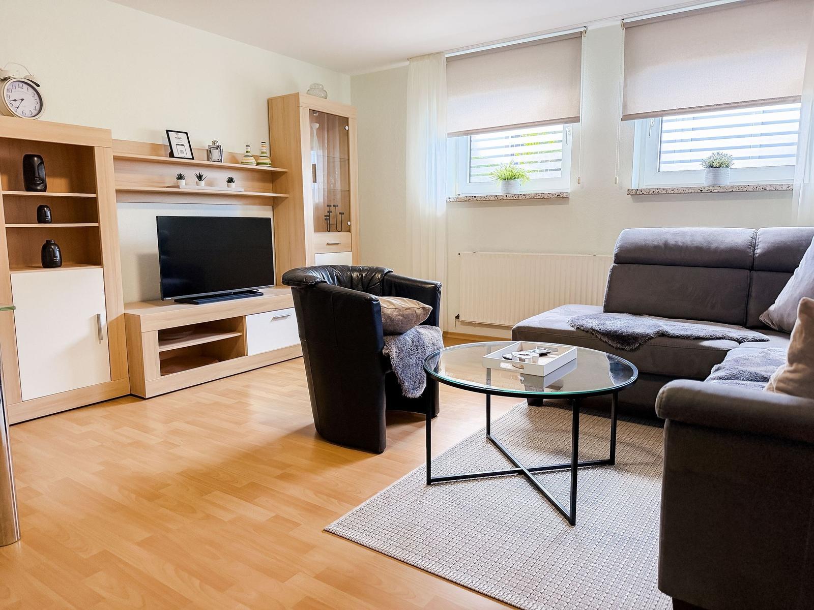 Wohnzimmer mit Sofa, Sessel, Glas-Tisch und Fernseher. Holzfußboden und Fenster mit Jalousien.