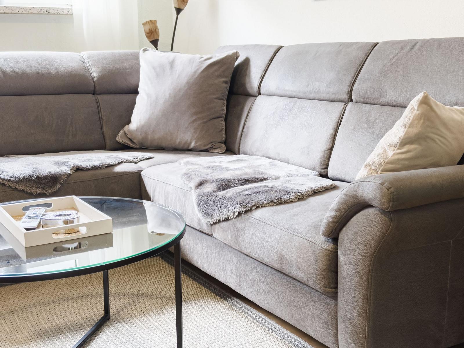 Ein grauer Sofa mit Kissen und Decke. Vorne steht ein runder Glas-Tisch.