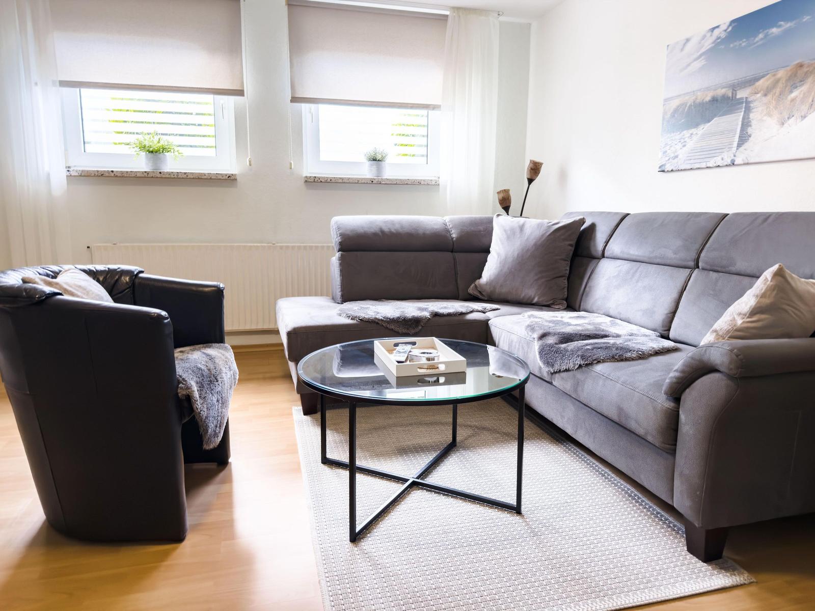 Ein Wohnzimmer mit grauem Sofa, schwarzer Ledercouch und runden Glas-Tisch.