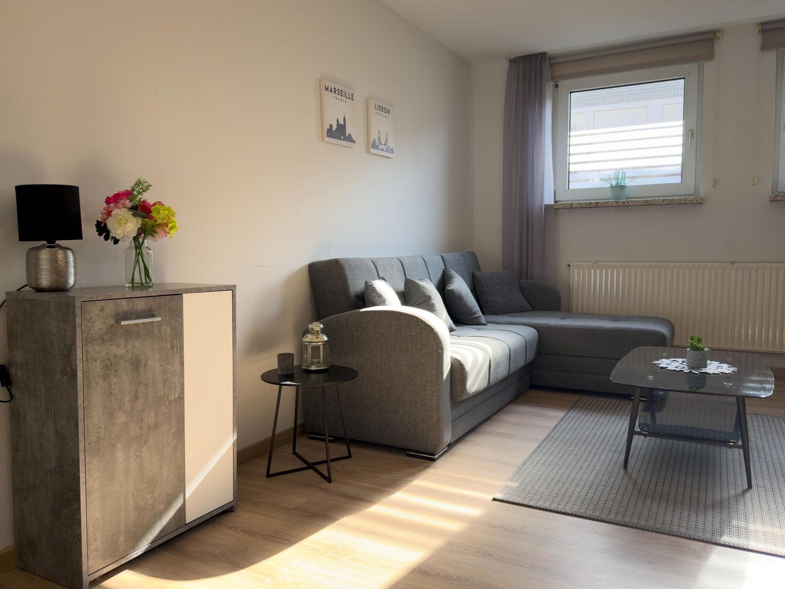 Wohnzimmer mit grauem Sofa, Tisch und Schrank. Fenster mit Jalousie und Blumen.