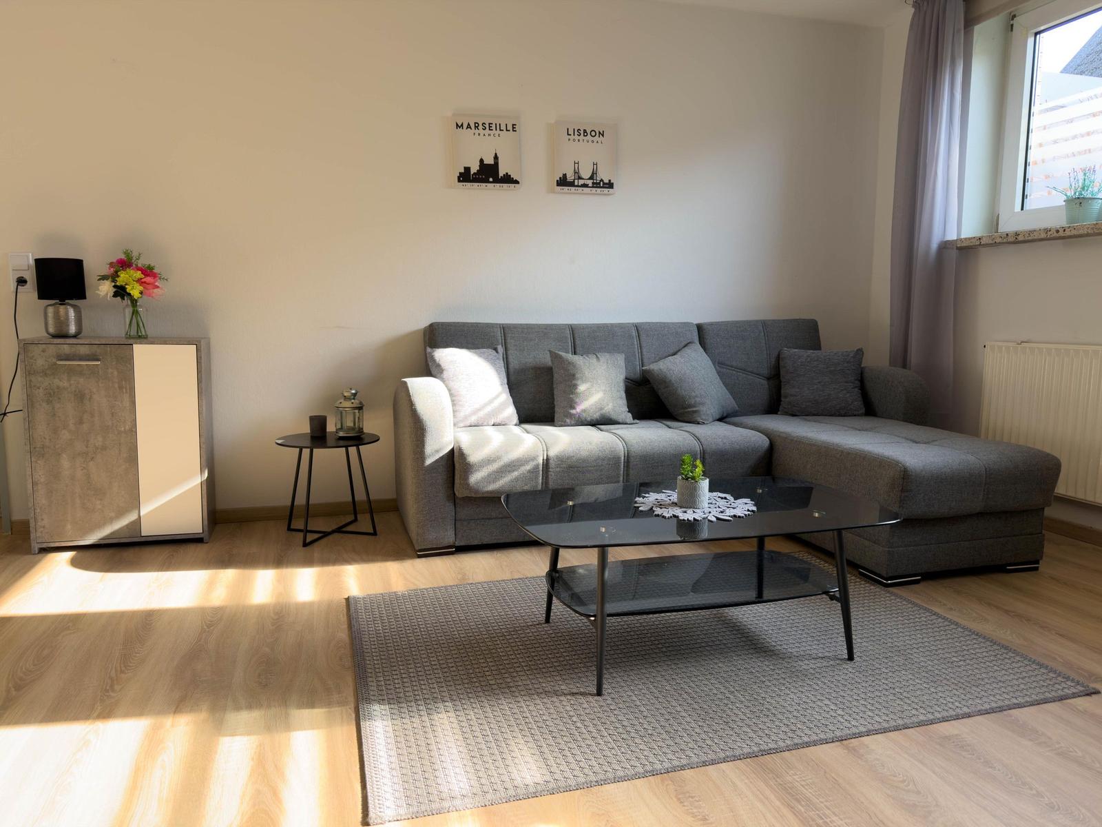 Wohnzimmer mit grauem Sofa, Glas-Tisch und Holzboden. Wandbilder von Marseille und Lissabon.