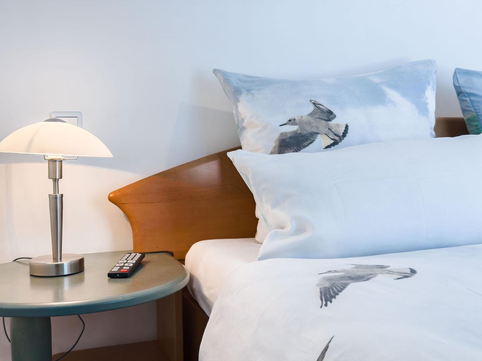A bed with pillows and bedding featuring seagull patterns. Nightstand with lamp and remote control.