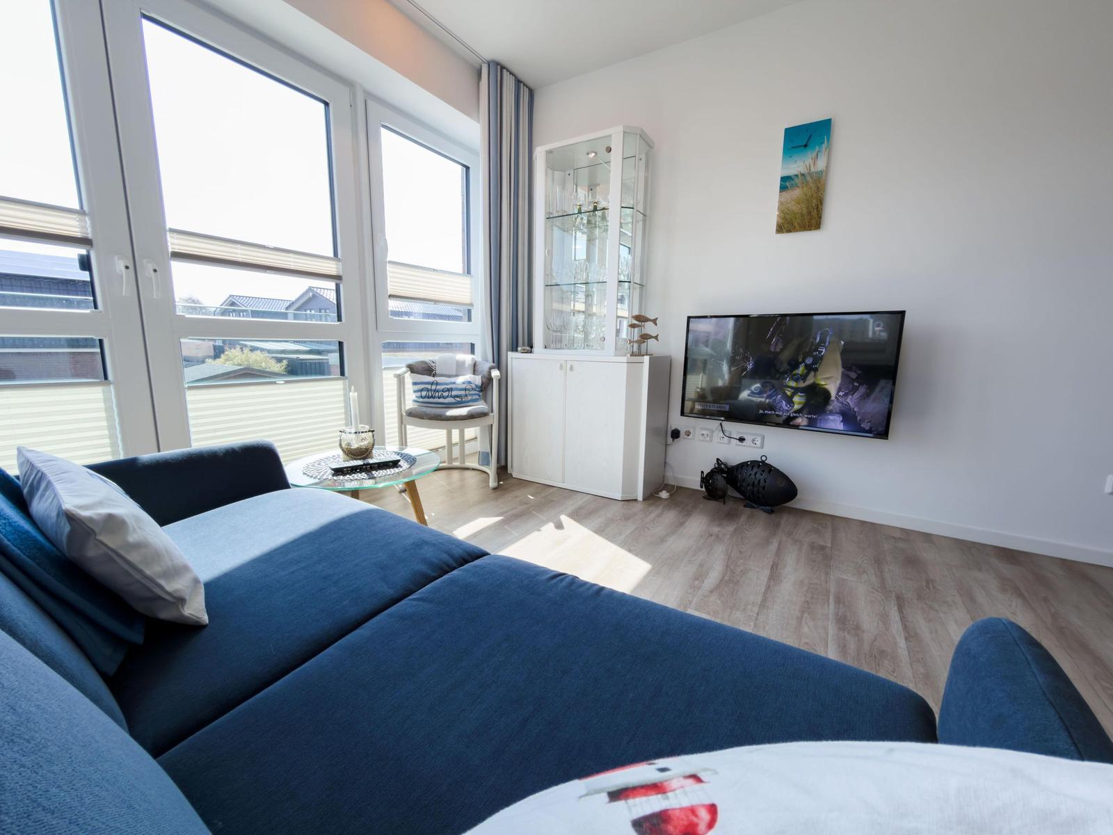 A living room with a blue sofa, large windows, and a TV mounted on the wall.