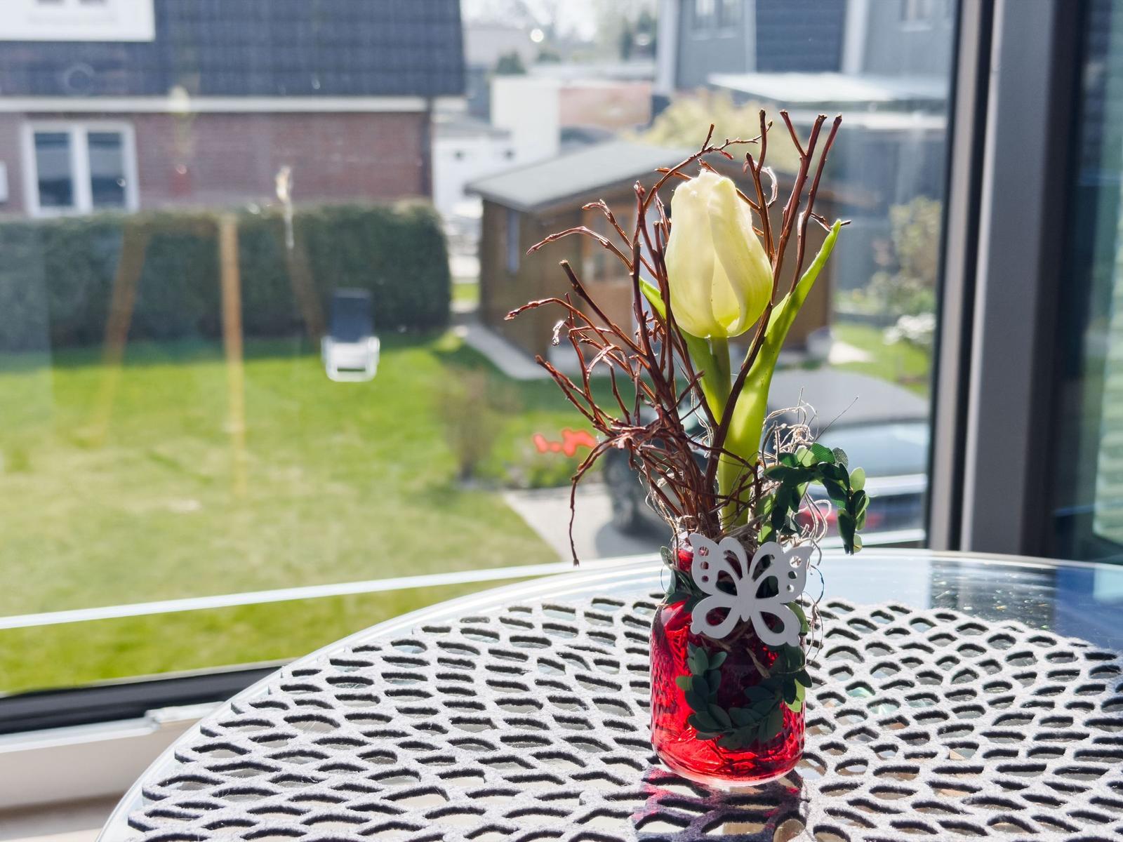 A tulip in a red glass vase sits on a table by a window overlooking a garden.