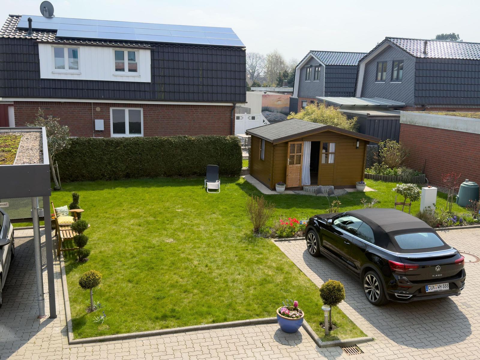 Backyard with garden, wooden shed, and paved parking area with car.
