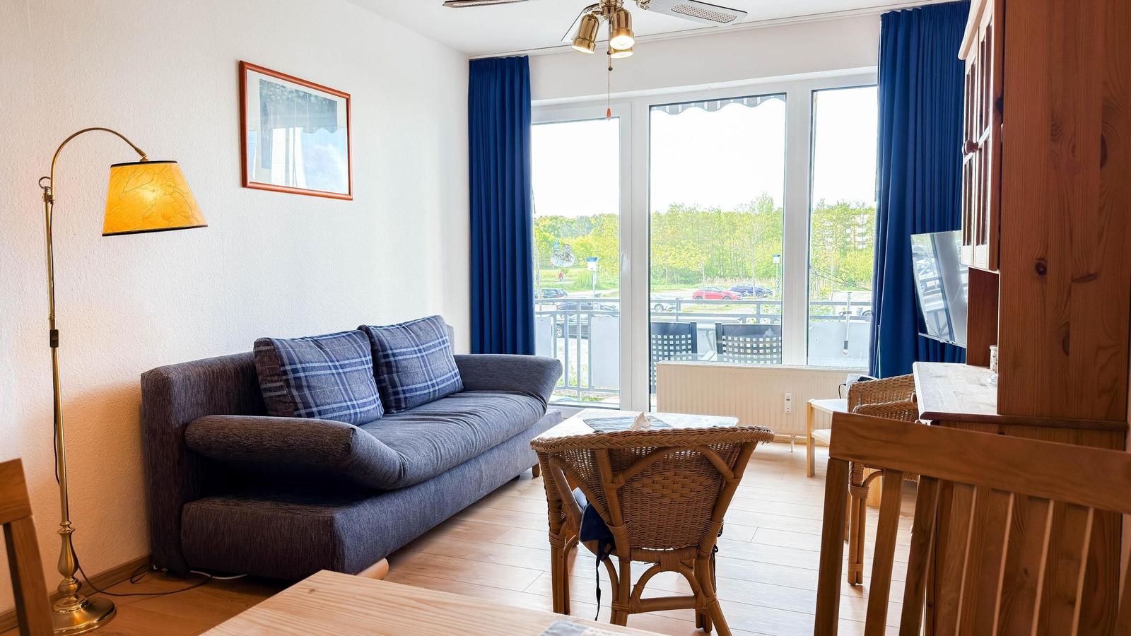 Wohnzimmer mit Sofa, Tisch und Stühlen. Große Fenster mit Blick auf Balkon und Straße.