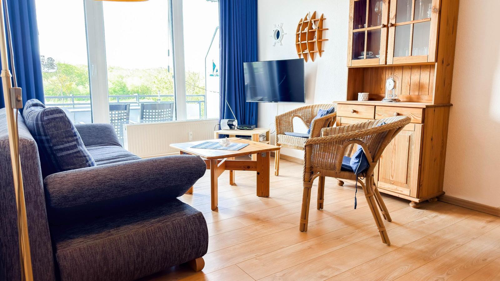 Wohnzimmer mit Sofa, Esstisch, Fernseher und Holzmöbel. Blick auf Balkon mit Garten.