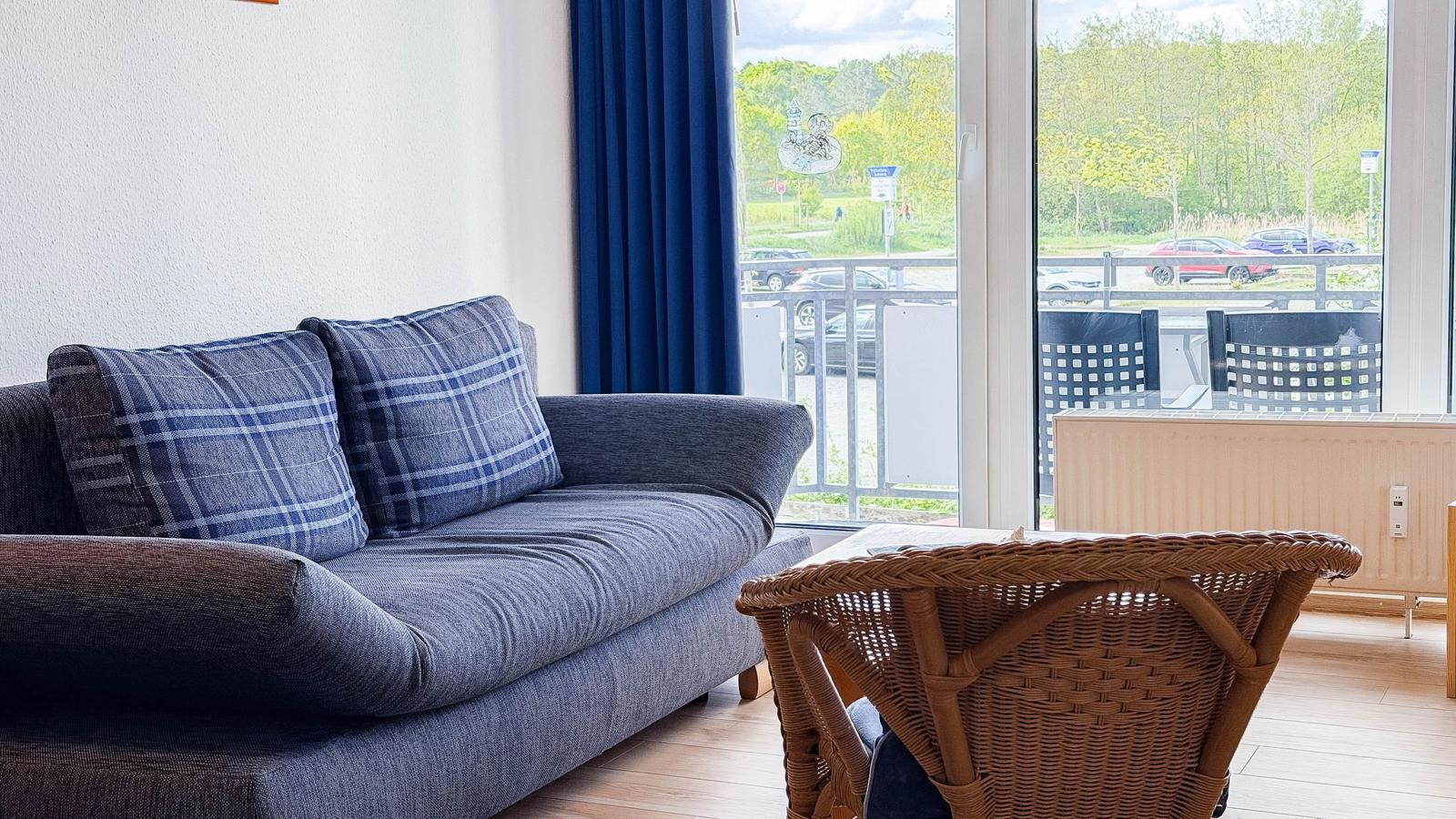 Ein Wohnzimmer mit Sofa, Kissen und Stuhl. Durch das Fenster ist eine Terrasse und Bäume sichtbar.