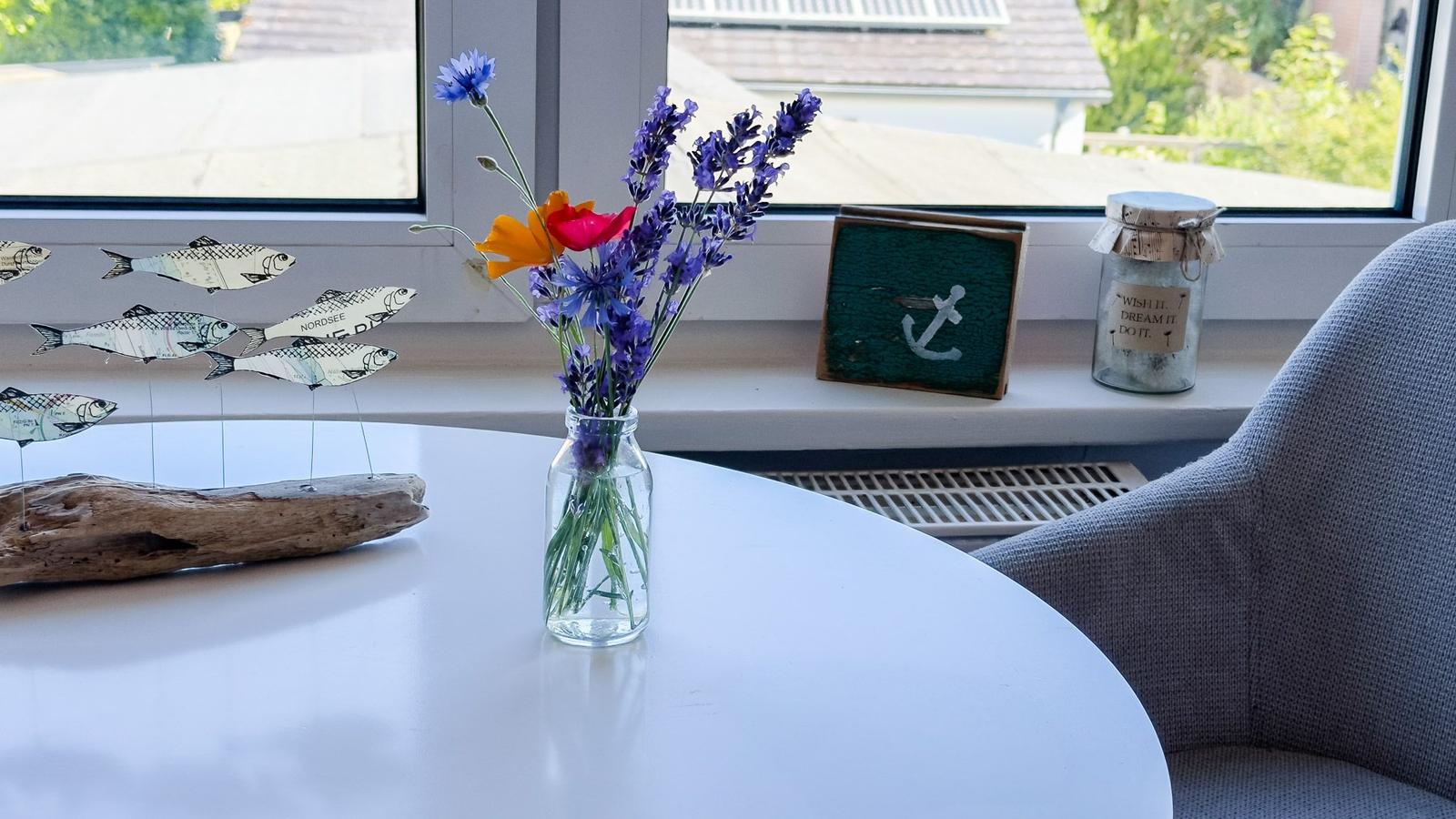 Auf dem Tisch steht eine Vase mit Blumen. Im Hintergrund sind Fenster und ein Sessel zu sehen.