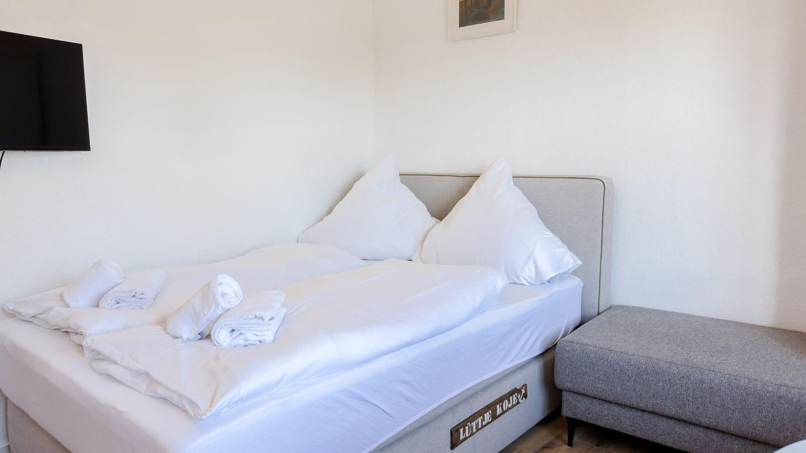 Ein Bett mit weißen Laken und Kissen. Daneben steht ein grauer Sitzhocker.