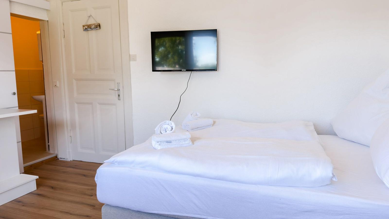 Schlafzimmer mit weißem Bett, Fernseher an der Wand und Tür zum Bad.