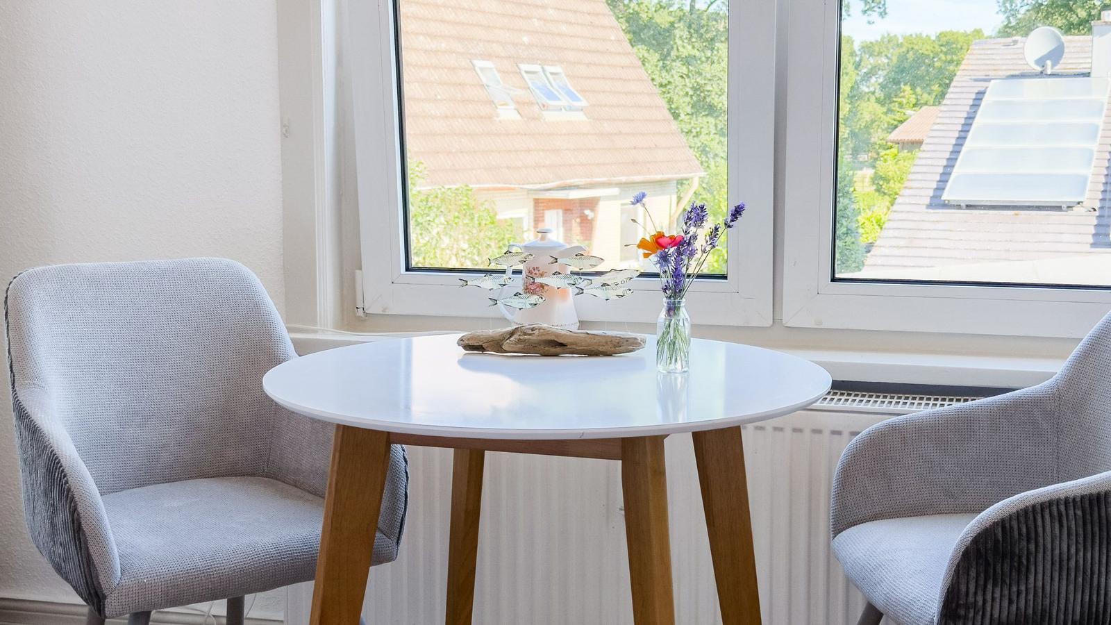 Rundtisch mit zwei Stühlen vor Fenster mit Blick auf Dächer und Grün.