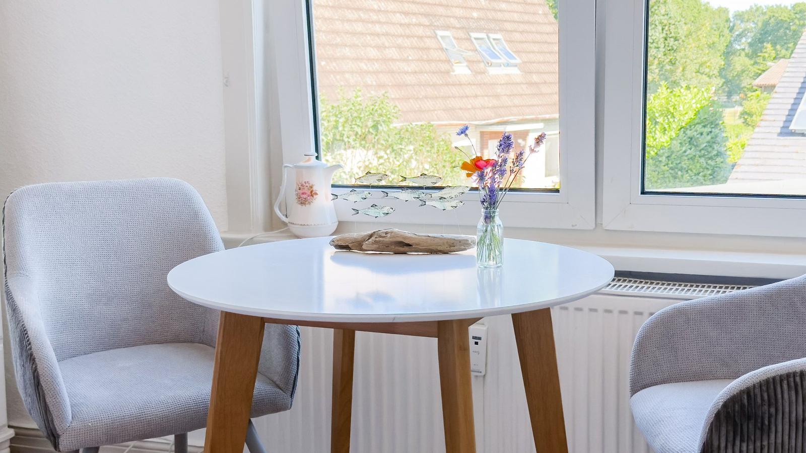 Rundes Tisch mit Stühlen vor Fenster, Blumen und Teekanne darauf.