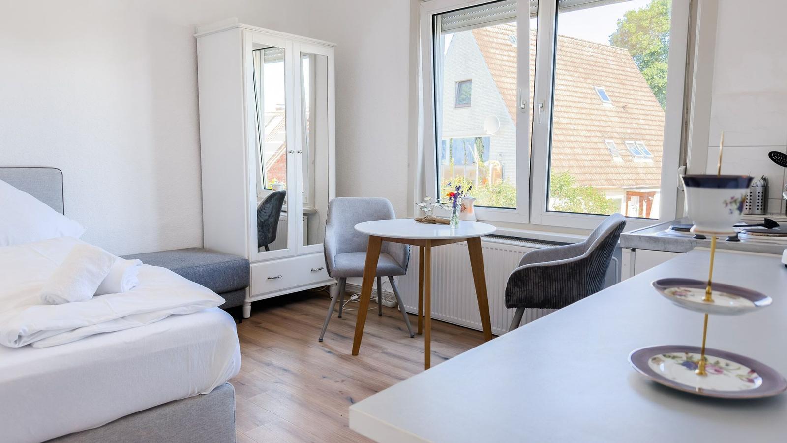 Schlafzimmer mit Bett, Kleiderschrank und Tisch. Fenster zeigt Dach und Grün.