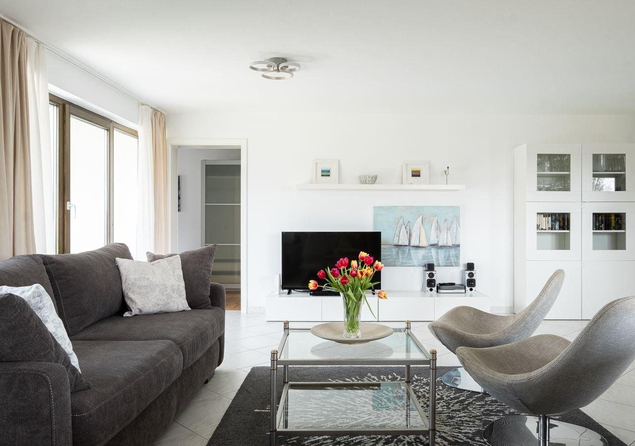 Wohnzimmer mit Sofa, Glas-Tisch und zwei Stühlen. Fernseher und Wandbild sind sichtbar.