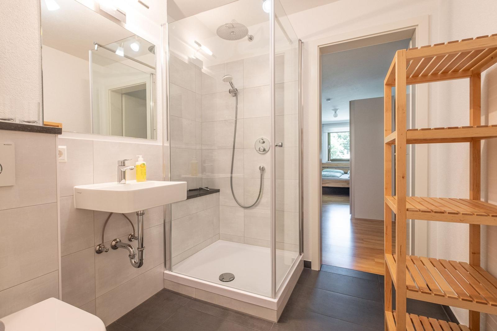 Badezimmer mit Dusche, Waschbecken und Holzregal.