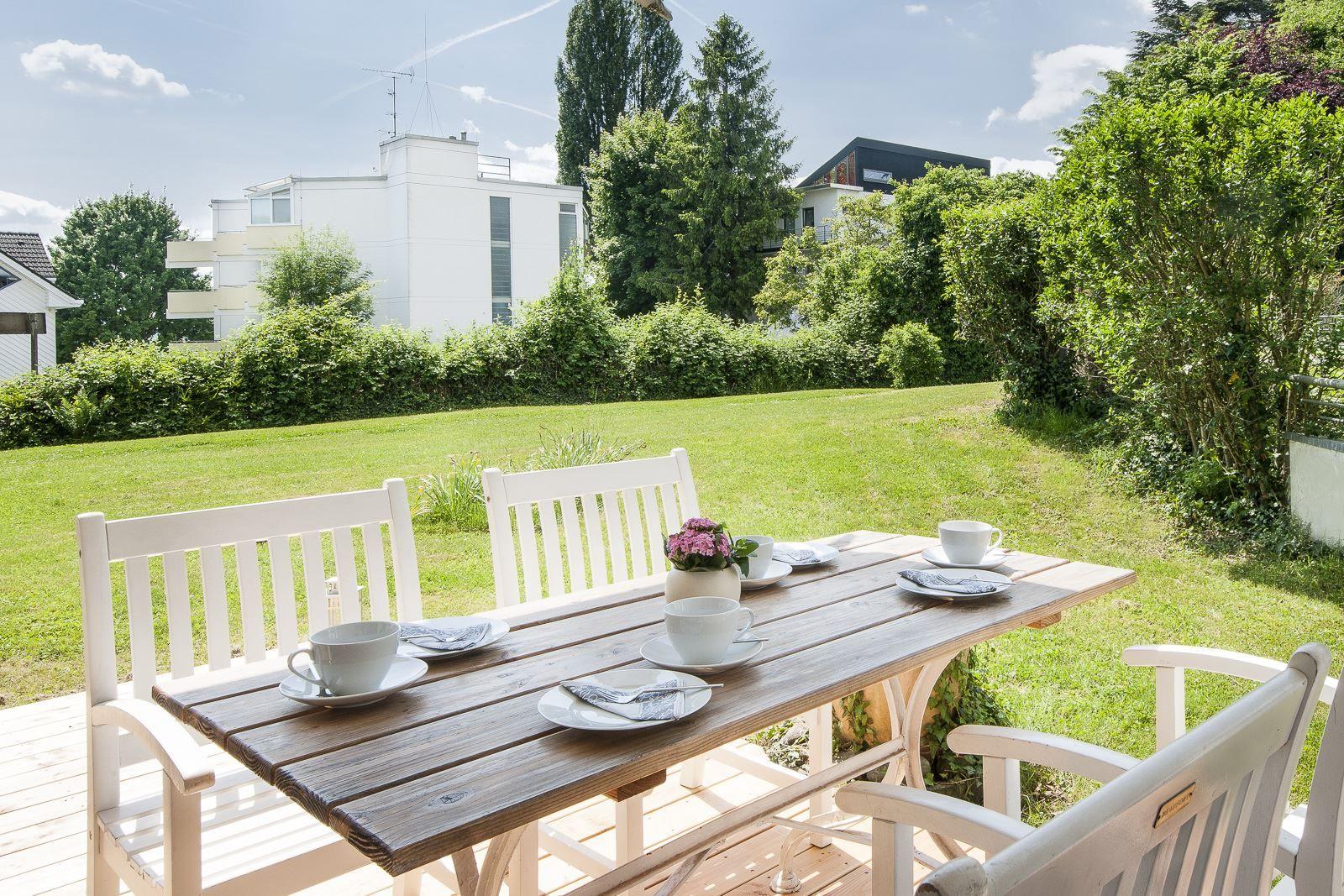 Holztisch mit weißen Stühlen auf Terrasse, Blick auf grünen Rasen und Bäume.