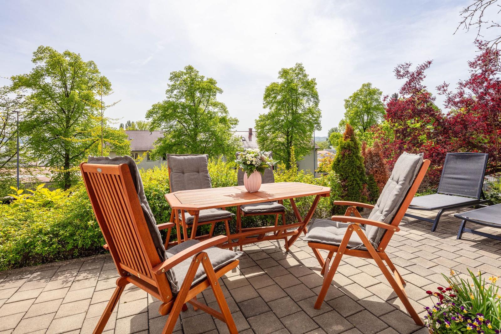 Terrasse mit Holztisch, Stühlen und Liegen. Umgeben von grünen Bäumen und Sträuchern.