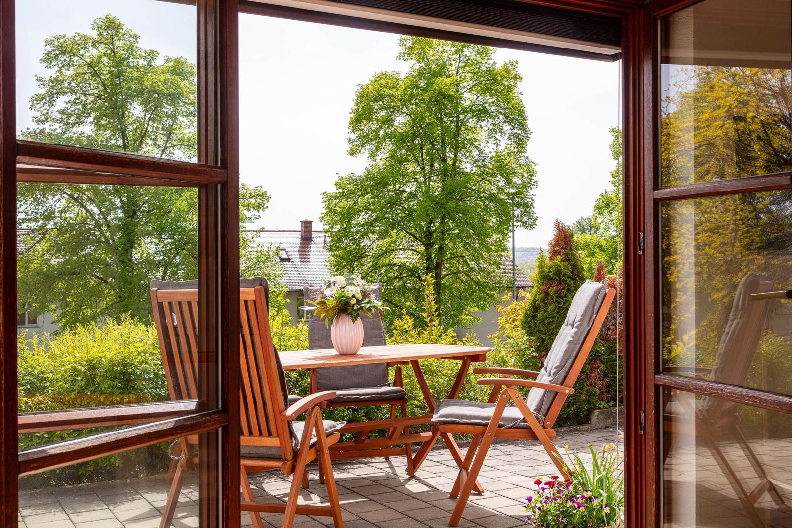 Terrasse mit Holztisch, Stühlen und Blumenarrangement. Blick auf grüne Bäume und Garten.